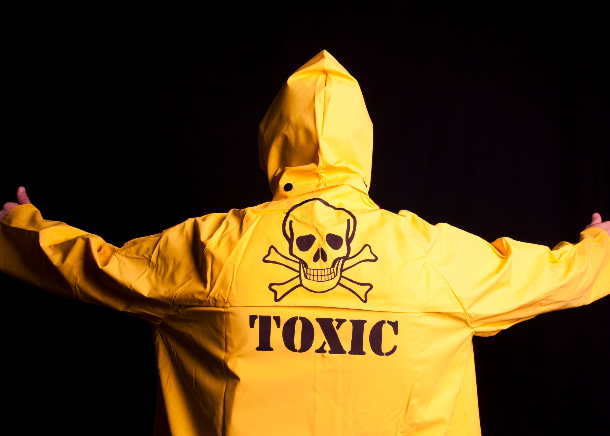 Image of the back of a person wearing a yellow rain jacket with the words toxic under the skull and bones symbol for poison.