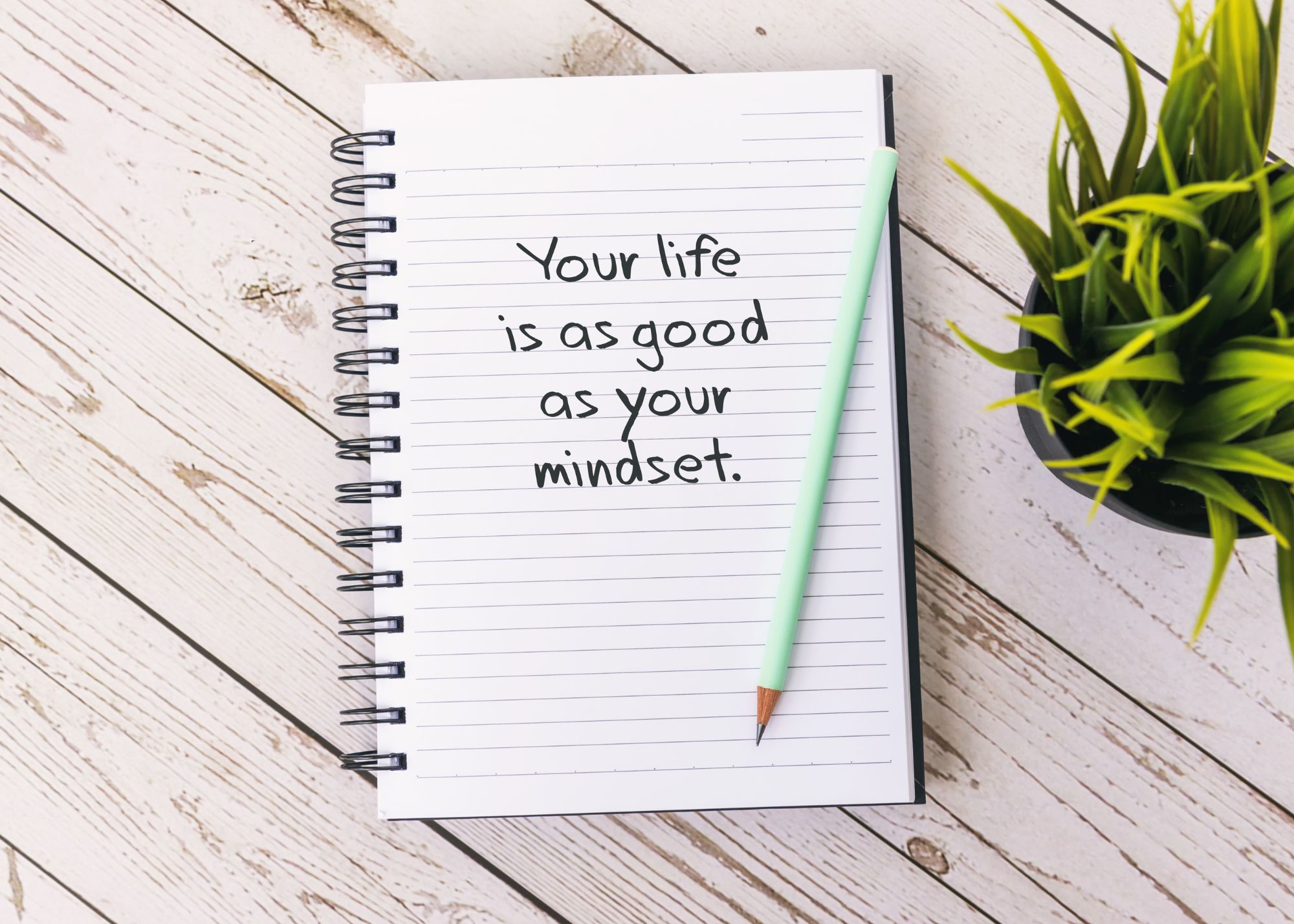 A notebook on a white desk with a message written on the page that says, "Your life is as good as your mindset." An aqua-colored pencil is laid across the notebook, which is on the desk next to a plant.