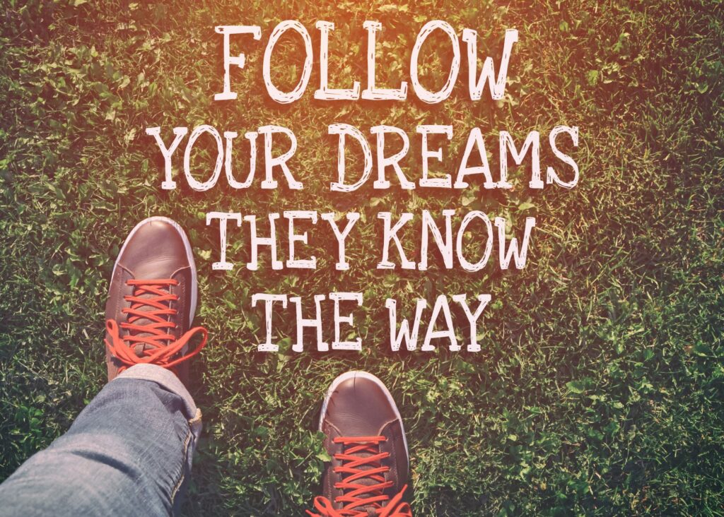 Image of an individuals shoes with red laces standing on grass. A message with white letters overlays the grass that reads, "Follow your dreams, they know the way."