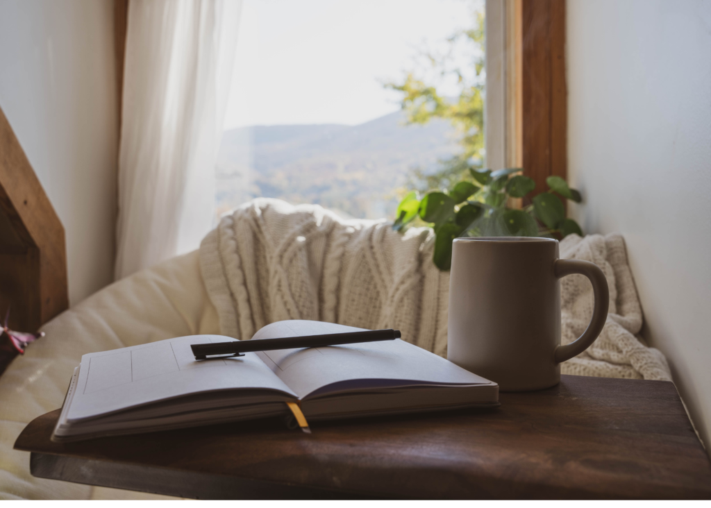 A journal is open to blank pages on a wooden table with a pen laying across it. A clay mug is to the right of the journal on the table. In the back ground you see a cozy thick knit ivory blanket dropped over the back of a chair, which is in front of a window with ivory drapes and a plant in the window sill. The image portrays the intersection of warmth of connections and mindfulness.