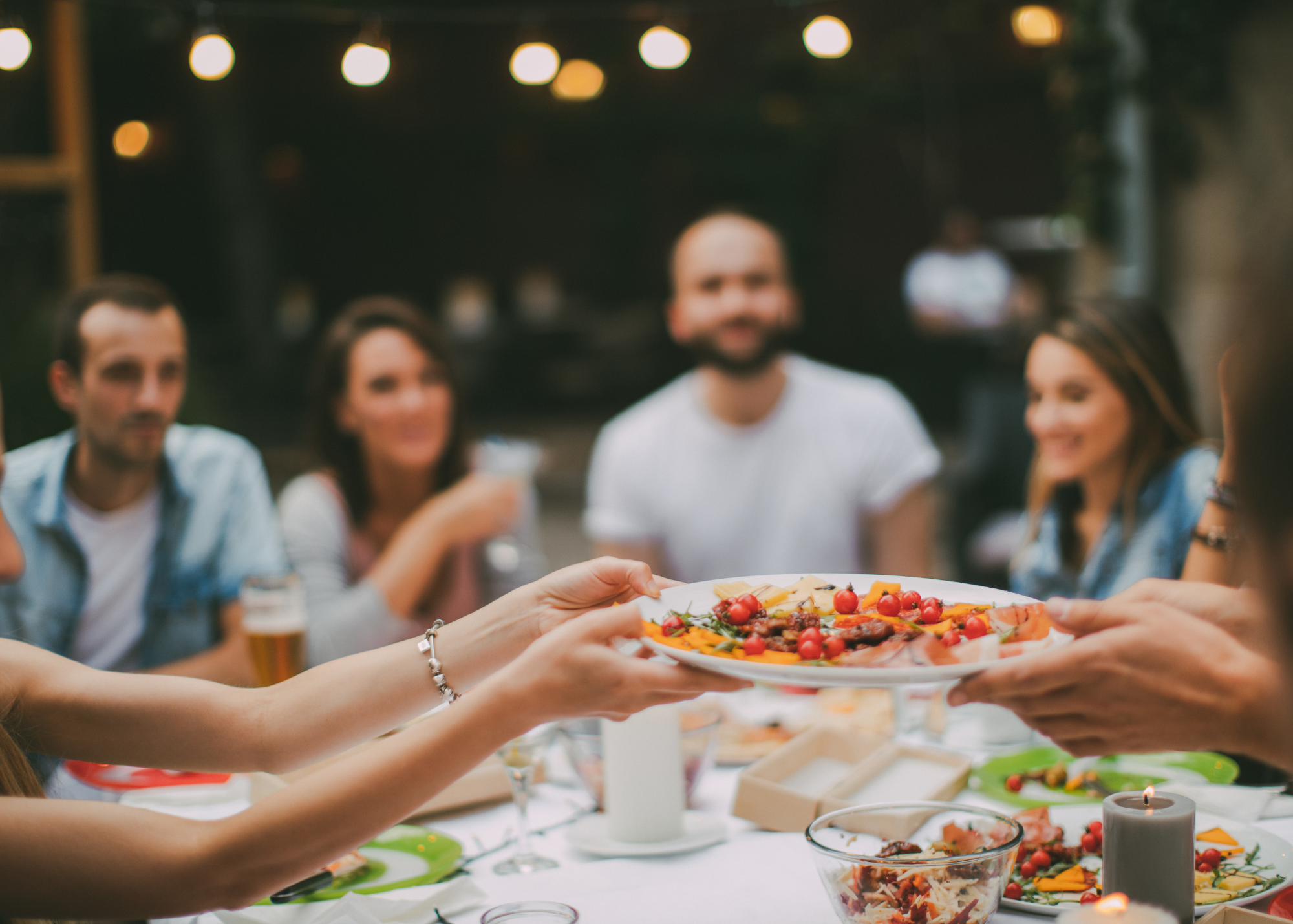 A blurry background of smiling loved ones around a table reaching out and sharing an in-focus pizza in the foreground. The image demonstrates warmth of connections and mindfulness.